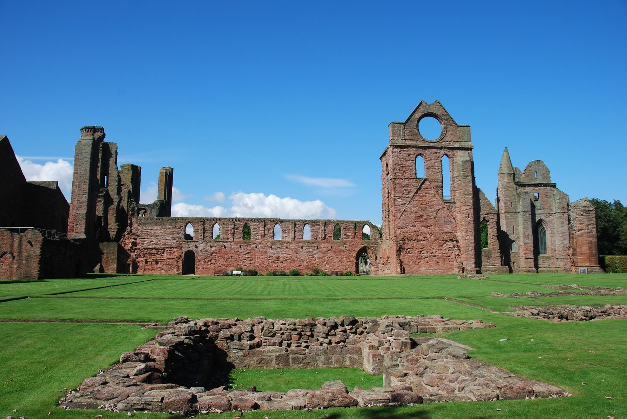 Ruined red-stone arches stand on grassy coastal grounds overlooking the sea beneath a cloudy blue sky in Arbroath.