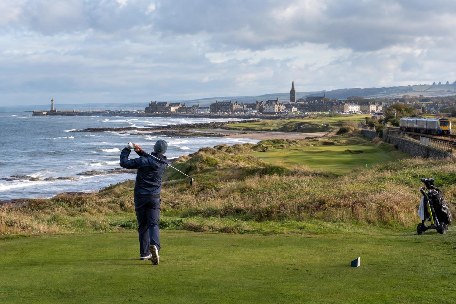 A golfer swings on a coastal course in Angus, with waves, a seaside town and a passing train beneath cloudy skies.